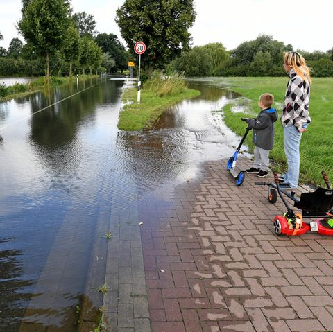 Eine Frau und ein Junge mit einem Roller stehen auf einem Fußweg. Beide schauen auf eine Landstraße, die überflutet ist