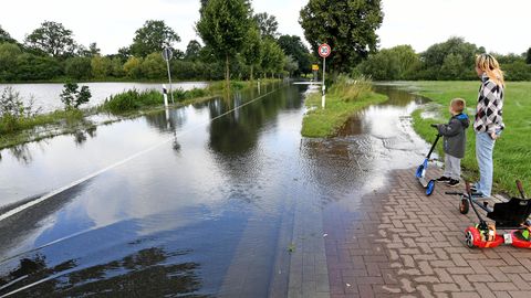 Eine Frau und ein Junge mit einem Roller stehen auf einem Fußweg. Beide schauen auf eine Landstraße, die überflutet ist