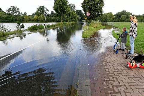 Eine Frau und ein Junge mit einem Roller stehen auf einem Fußweg. Beide schauen auf eine Landstraße, die überflutet ist