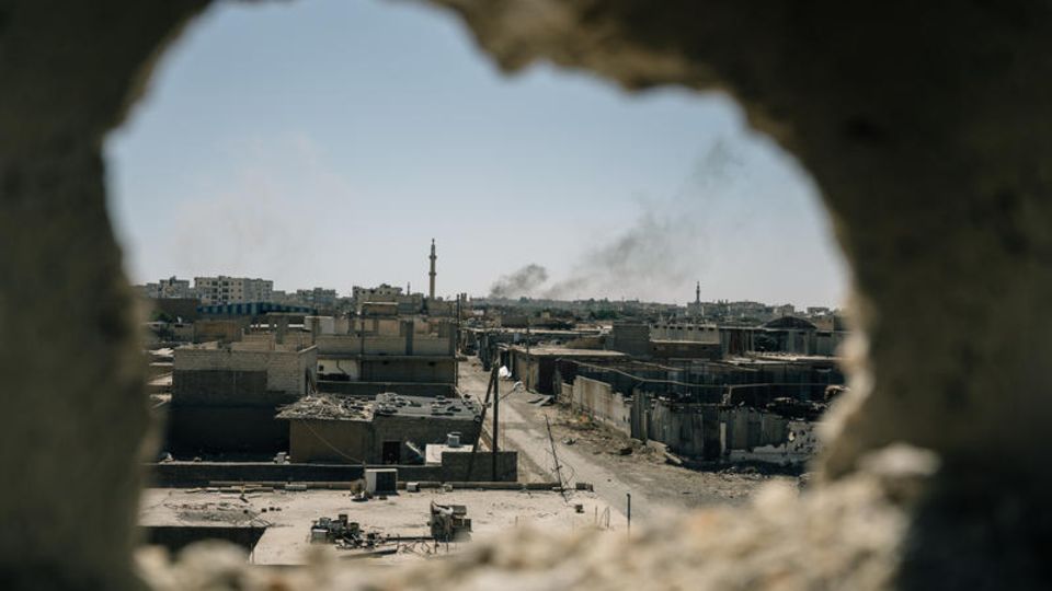 Der Blick durch ein zerstörtes Haus auf die Altstadt von Raqqa. In der Ferne steigt Rauch von einem Bombenangriff auf