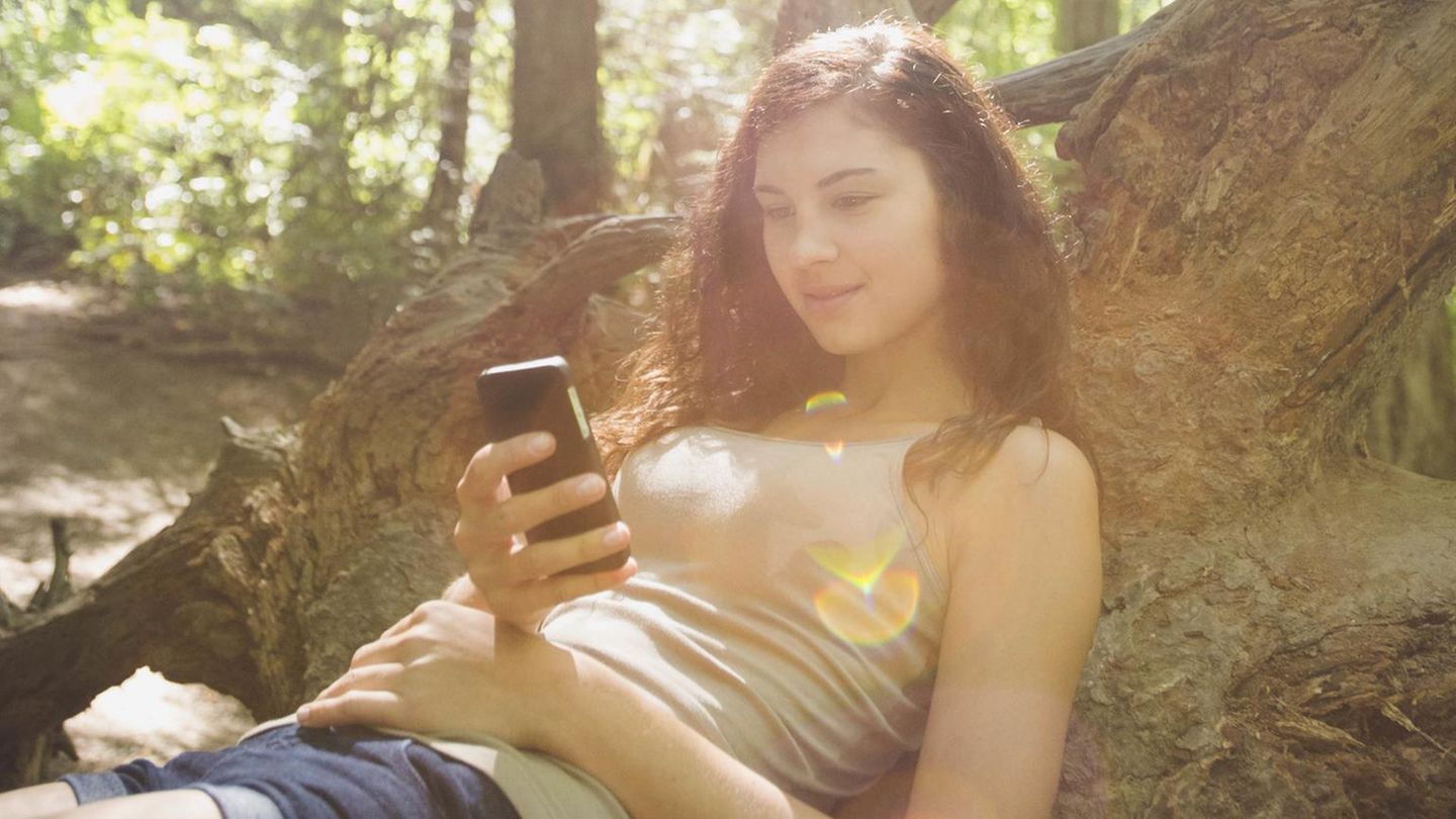 Eine junge Frau hat ihr Smartphone in der Hand und sitzt im Wald