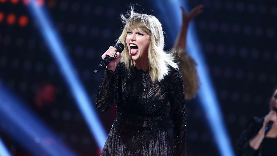 Taylor Swift steht in einem schwarzen, kurzen Kleid mit Fransen auf der Bühne und singt in ein Mikrofon