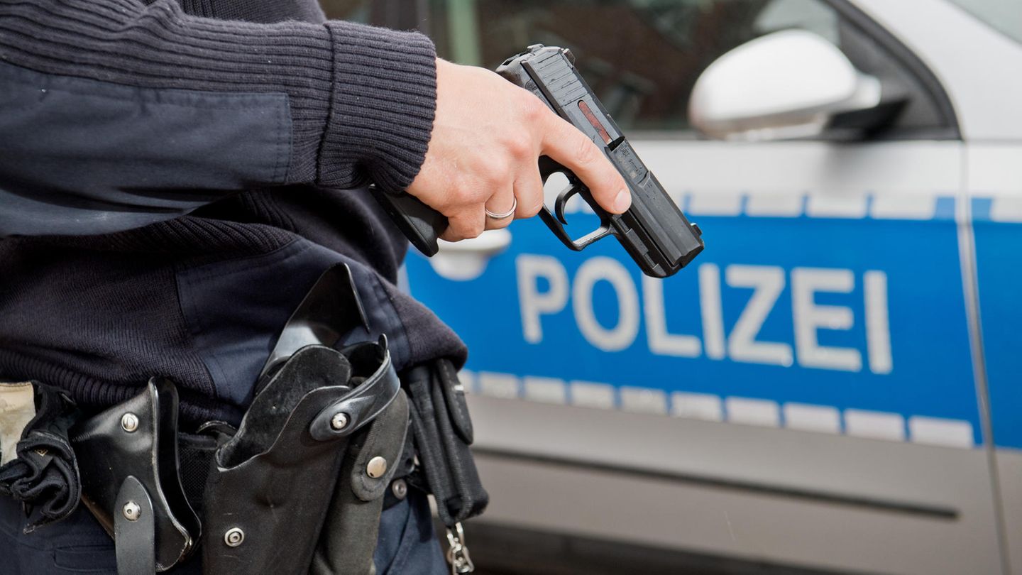 Ein Polizist hält seine Dienstwaffe vor einem Polizeiwagen (Symbolbild)