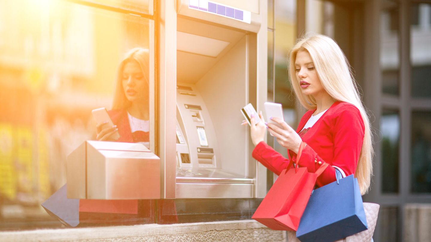 Online-Banking Eine junge Frau steht mit Taschen beladen vor einem Geldautomat und schaut auf das Smartphone