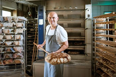 Bäcker Arnd Erbel steht in seiner Backstube und zeigt ein Brot, dass er gerade aus dem Ofen gezogen hat