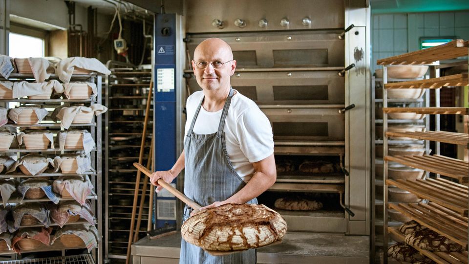 Bäcker Arnd Erbel steht in seiner Backstube und zeigt ein Brot, dass er gerade aus dem Ofen gezogen hat