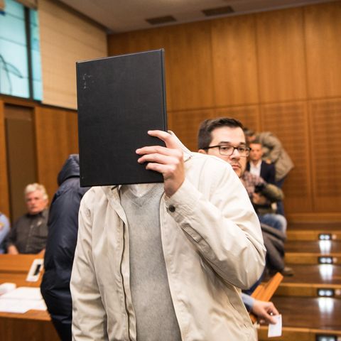 Ein Mann in heller Windjacke und grauem Pullover hält sich im Landgericht Frankfurt eine schwarze Mappe vor das Gesicht.