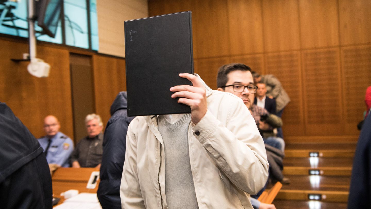 Ein Mann in heller Windjacke und grauem Pullover hält sich im Landgericht Frankfurt eine schwarze Mappe vor das Gesicht.