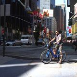 Broadway – Times Square  Ich hatte Angst vor dem Broadway, aber ich merke gleich: Der Broadway ist mein Freund. Ein dicker Fahrradstreifen säumt den ehemaligen Indianerpfad, der Manhattan diagonal durchschneidet. Ich gewinne an Sicherheit, verschmelze mit dem Metropolenwahnsinn um mich herum. Ich beobachte die radfahrenden New Yorker, imitiere sie. Bei Rot über die Fahrradampel, auch wenn dort ein Polizeiauto parkt? No problem. Fahren auf dem Bürgersteig? No way! Das Menschengewusel wird dichter, das neonfarbene Reklameblinken schriller, ich umkurve einen Kran, der den Fahrradweg blockiert, dann lande ich in der totalen Reizüberflutung. Der Duft des heißen Wurstwassers der Hot-Dog-Stände vermischt sich mit dem gerösteter Mandeln und der muffigen Luft der U-Bahn-Schächte. Micky Maus winkt mir zu. Eine Freiheitsstatue will mich umarmen. Die verkleideten Straßenkünstler geben sich alle Mühe, den Touristen das Kleingeld zu entlocken. Mein Klingeln geht im Großstadtlärm unter. Ich gebe auf und steige ab. Times Square. Surreal, wie eine Comicverfilmung der Wirklichkeit. Ich schiebe das Fahrrad einmal quer durch die Menschenmenge – und bin auf schätzungsweise 150 Selfies gebannt. 