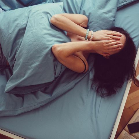 Eine Frau liegt angezogen im Bett und schlägt die Hände vors Gesicht