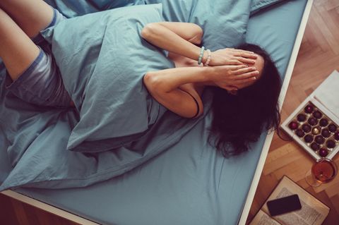 Eine Frau liegt angezogen im Bett und schlägt die Hände vors Gesicht