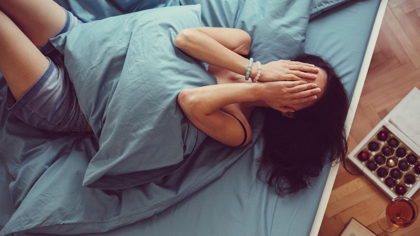 Eine Frau liegt angezogen im Bett und schlägt die Hände vors Gesicht