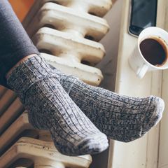 Die Füße einer Frau liegen in Wollsocken auf einer Heizung. Auf der Fensterbank stehen ein Kaffeebecher und ein Smartphone