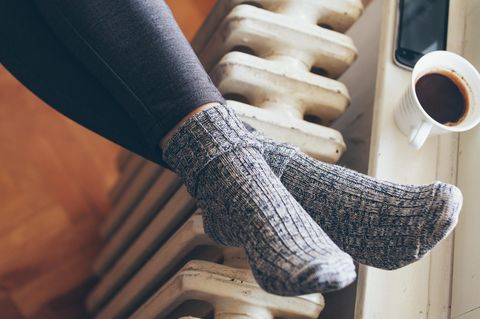 Die Füße einer Frau liegen in Wollsocken auf einer Heizung. Auf der Fensterbank stehen ein Kaffeebecher und ein Smartphone