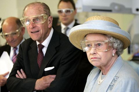 Queen Elizabeth II steht im himmelblauen Kostüm und oassendem Strohhut mit einer durchsichtigen Schutzbrille in einem Labor