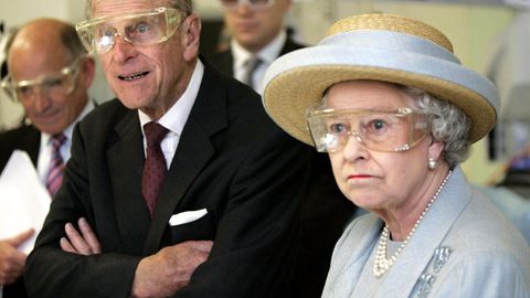 Queen Elizabeth II steht im himmelblauen Kostüm und oassendem Strohhut mit einer durchsichtigen Schutzbrille in einem Labor