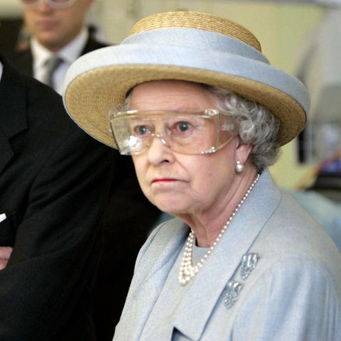 Queen Elizabeth II steht im himmelblauen Kostüm und oassendem Strohhut mit einer durchsichtigen Schutzbrille in einem Labor