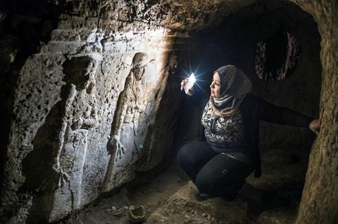 Im Schein ihres Handys hockt eine Frau mit Kopftuch vor einem steinernen Relief und betrachtet zwei gemeißelte Figuren