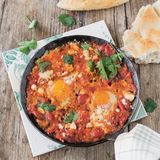 Shakshuka      Zutaten für 4 Personen:  2 Zwiebeln, 4 Knoblauchzehen, 2 rote Paprikaschoten, 1 TL Kreuzkümmelsamen, 1 TL Koriandersamen, 4 EL Olivenöl, 1 Dose Flaschentomaten (etwa 400 g), 1 Dose Cocktailtomaten (etwa 400 g), Salz, frisch gemahlener schwarzer Pfeffer, 1/2 Bund frischer Koriander, 4-8 Eier (pro Person 1-2 Eier), 150 g Feta    Außerdem: frisches Fladenbrot      Zubereitung:  Zwiebeln sowie Knoblauch abziehen und fein würfeln. Die Paprika waschen, trockentupfen, von Stielansätzen und Samen befreien und ebenfalls klein schneiden.  Die Kreuzkümmel- und Koriandersamen in einer Pfanne ohne Fett rösten, bis die Gewürze duften. Im Mörser fein zerreiben.  In einer großen oder zwei kleineren Pfannen das Olivenöl bei mittlerer Temperatur erhitzen. Die Zwiebeln und den Knoblauch darin anschwitzen, bis die Zwiebeln glasig sind. Die Paprika zugeben und 2-3 Minuten mitbraten. Die Tomaten in die Pfanne geben und mit dem Kochlöffel zerdrücken. Kräftig mit Salz, Pfeffer, gemahlenem Kreuzkümmel sowie Koriander würzen und die Mischung etwa 15 Minuten einköcheln lassen.   Den frischen Koriander waschen und trockenschütteln. Die Hälfte des Korianders hacken und unter die Tomaten rühren, von der anderen Hälfte die Blätter abzupfen und beiseitelegen.  Mit dem Kochlöffel Vertiefungen in die Sauce drücken und die Eier hineinschlagen. den Feta zerbröseln und auf den Tomaten um die Eier herum verteilen. Die Eier bei mittlerer Temperatur stocken lassen, bis das Eiweiß fest ist.  Salzen, pfeffern und mit Korianderblättern bestreut in der Pfanne servieren. Dazu passt frisches Fladenbrot. 