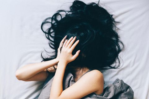 Eine Frau liegt im Bett und hält sich verzweifelt die Hände vor das Gesicht