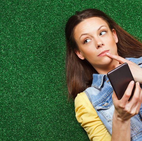Eine junge Frau liegt auf einer Wiese und denkt mit dem Smartphone in der Hand nach