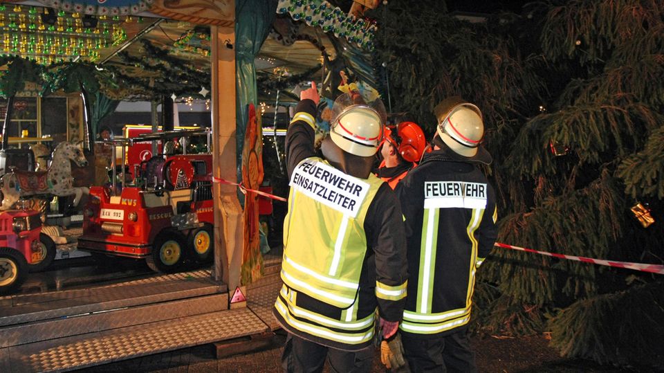 Nachrichten aus Deutschland: Zwei Feuerwehrmänner stehen vor dem zerstörten Kinderkarussell in Eschweiler bei Aachen