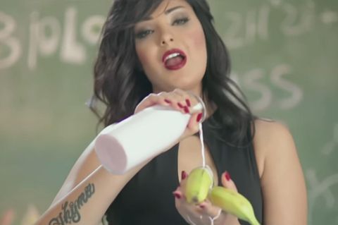 Die schwarzhaarige Sängerin Shyma steht vor einer Schultafel und gießt aus einer Flasche Milch über Bananen in ihrer linken Hand