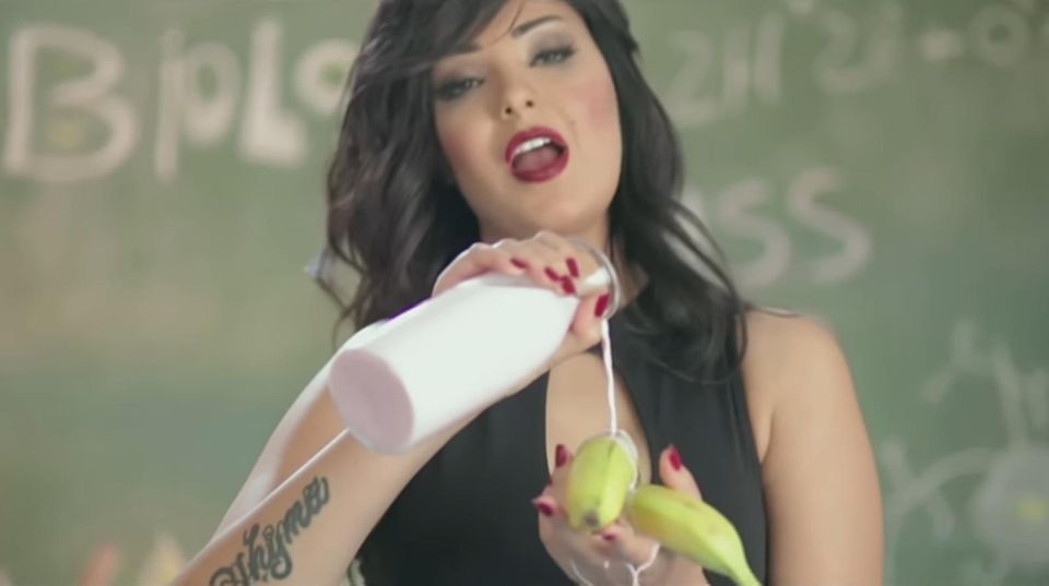 Die schwarzhaarige Sängerin Shyma steht vor einer Schultafel und gießt aus einer Flasche Milch über Bananen in ihrer linken Hand