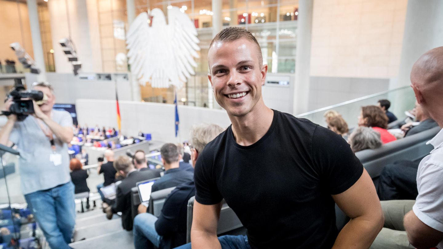 Ein muskulöser blonder Mann sitzt lächelnd in Jeans und T-Shirt auf der Zuschauertribüne im Bundestag