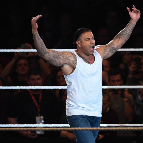 Tim Wiese steht in Jeans und weißem Tanktop im Wrestling-Ring und hebt herausfordern beide Arme