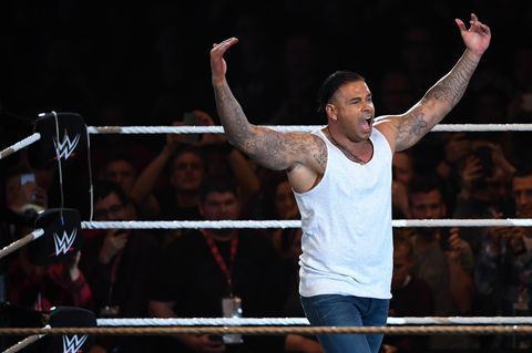 Tim Wiese steht in Jeans und weißem Tanktop im Wrestling-Ring und hebt herausfordern beide Arme