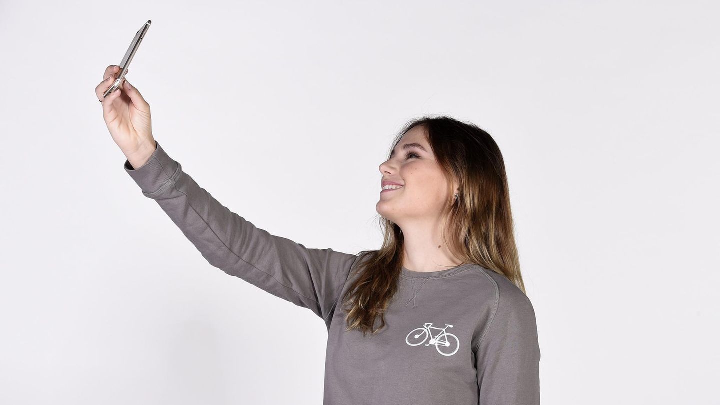Eine junge Frau in grauem Sweatshirt und mit halblangen Haaren macht mit einem Smartphone in der rechten Hand ein Selfie