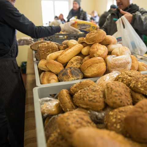 Eine Mitarbeiterin einer Tafel reicht einem Mann über Körbe mit Brötchen hinweg ein Brot