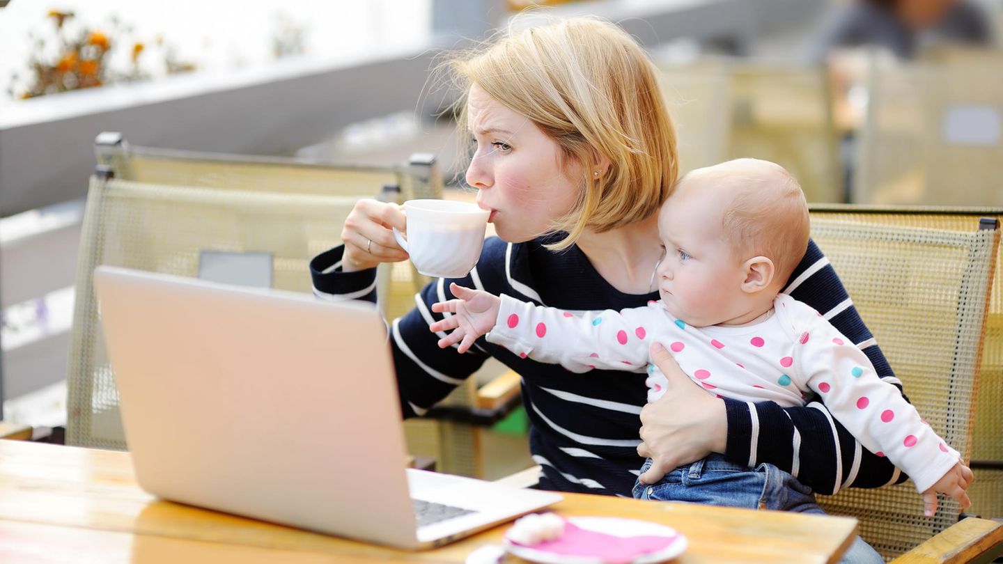 Eine Mutter mit Baby auf dem Schoß versucht, eine Tasse Kaffee zu trinken
