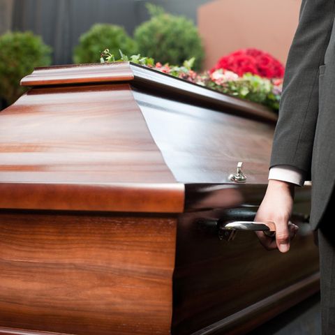 Sarg Bestatter Sargträger Neben einem braunen Holzsarg steht ein Mann in Anzug und hat in der rechten Hand einen Griff des Sarges