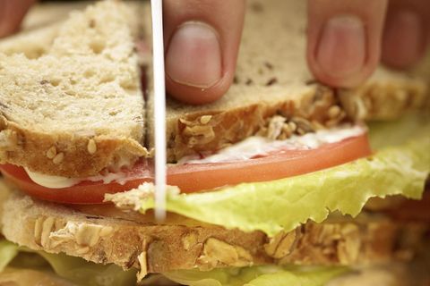Auf einem Holzbrett liegt ein Sandwich. Vier Finger halten das Sandwich, während eine Messerklinge es durchschneidet.