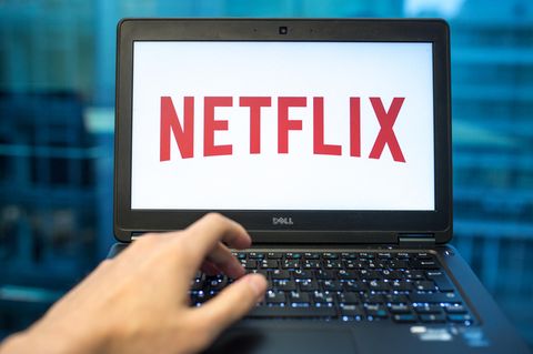 Ein Laptop-Bildschirm zeigt auf weißem Grund den roten "Netflix"-Schriftzug. Eine linke Hand liegt auf der Tastatur