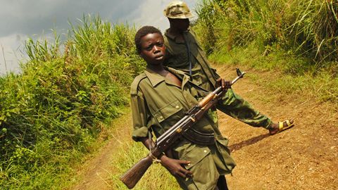 Ein schwarzer Junge in Uniform und mit einer AK47 steht auf einem Feldweg