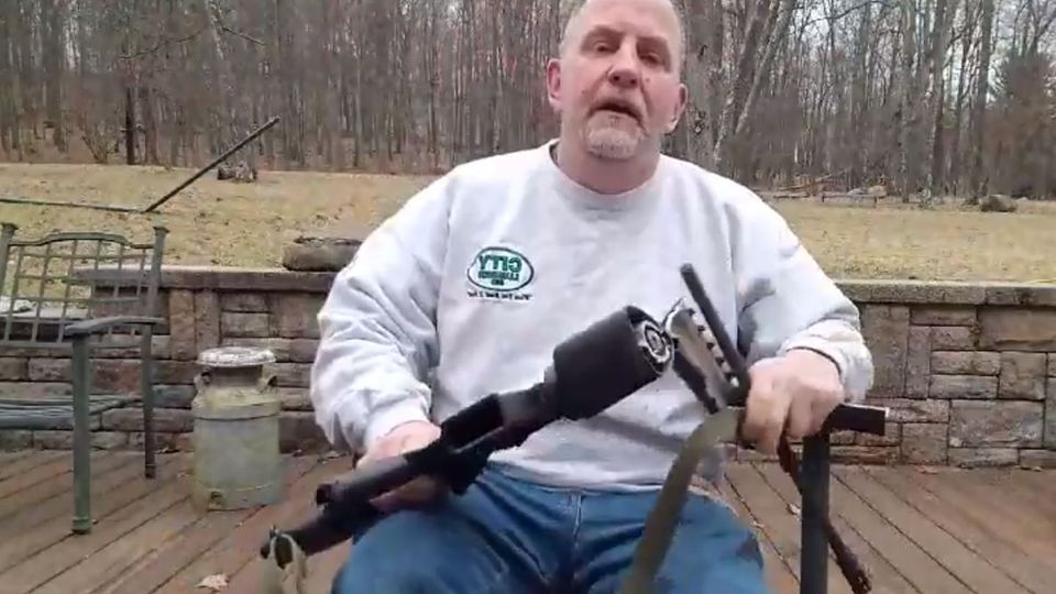 Ein älterer Mann mit Jeans und grauem Sweatshirt sitzt mit seiner zersägten AR-15 in den Händen auf einer Terrasse