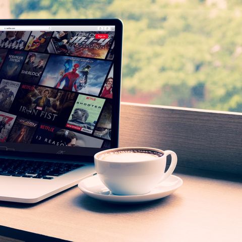 Eine Tasse Kaffee steht neben einem Laptop, auf dem das Streaming-Angebot von Netflix geöffnet ist.