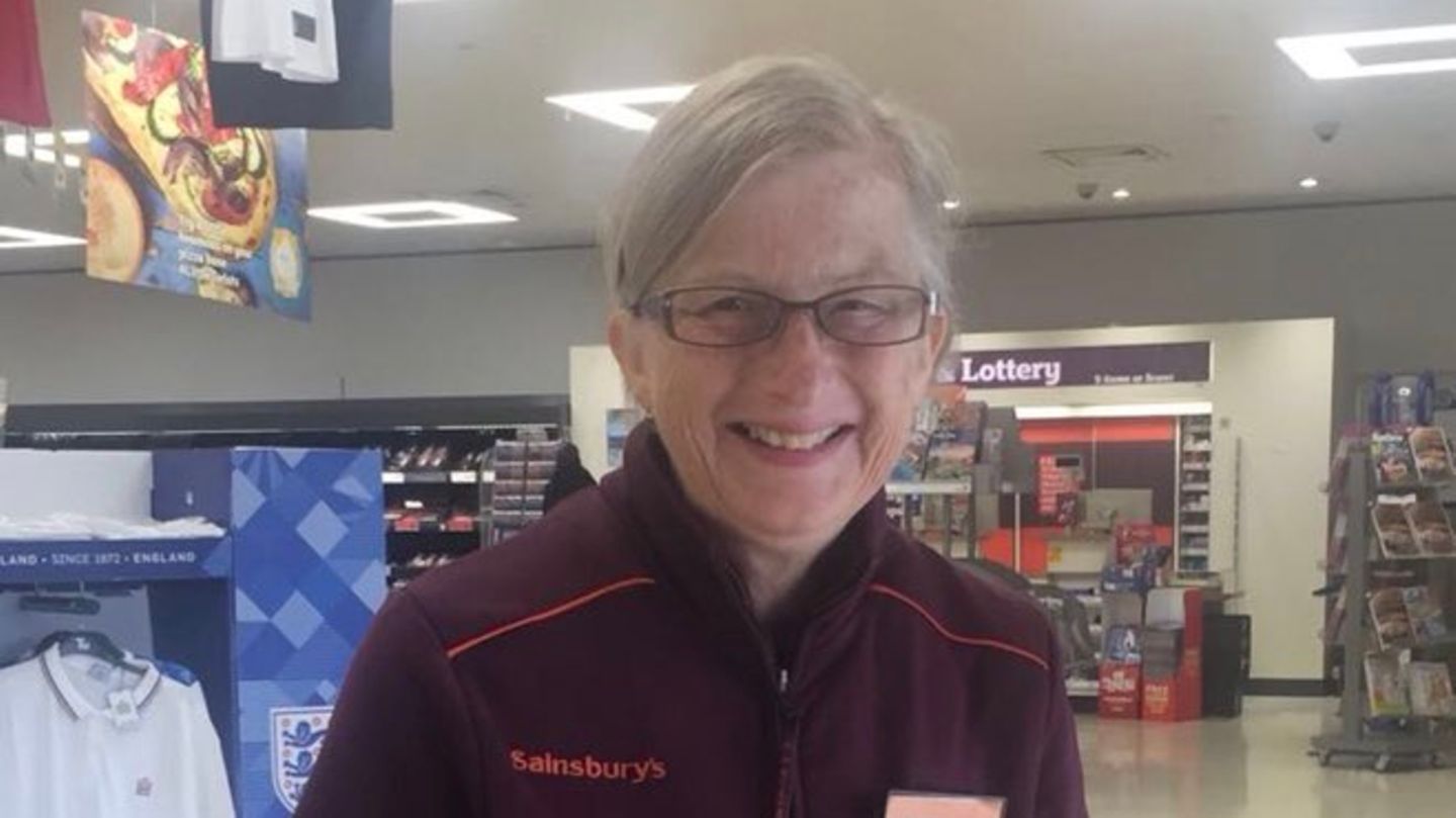 Eine grauhaarige Frau mit Brille lächelt glücklich in die Kamera. Sie ist in einem Sainsbury's Supermarkt.