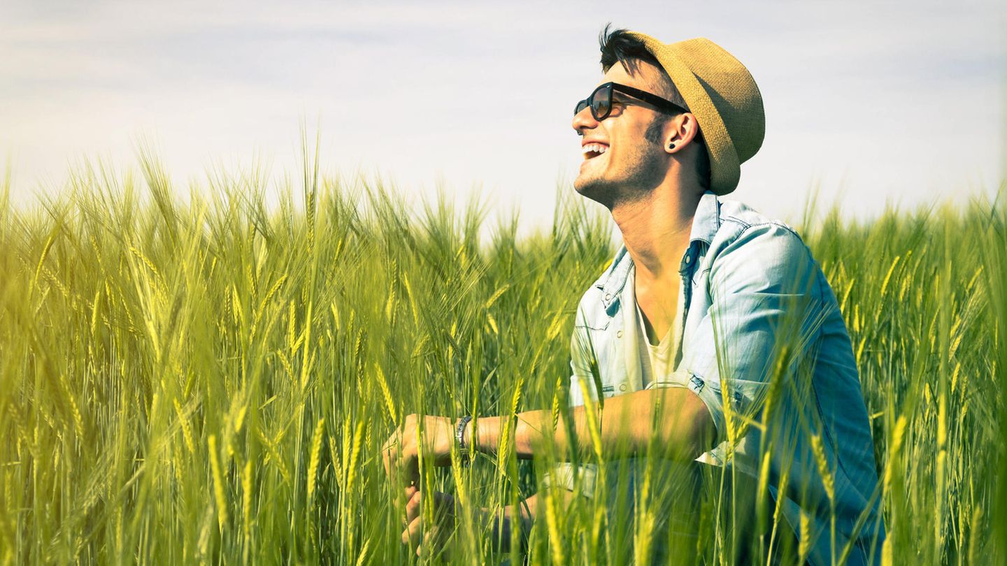 Junger Mann mit Sonnenbrille und Hut sitzt im Feld und lacht in die Sonne