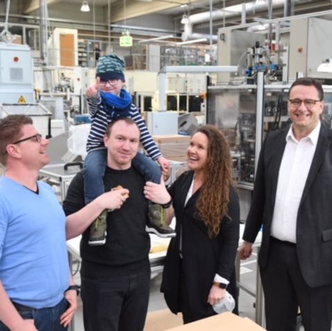 Drei Männer und eine Frau stehen lachend in einer Fabrik. Ein Mann trägt ein Kind auf der Schulter, das den Daumen hoch zeigt.