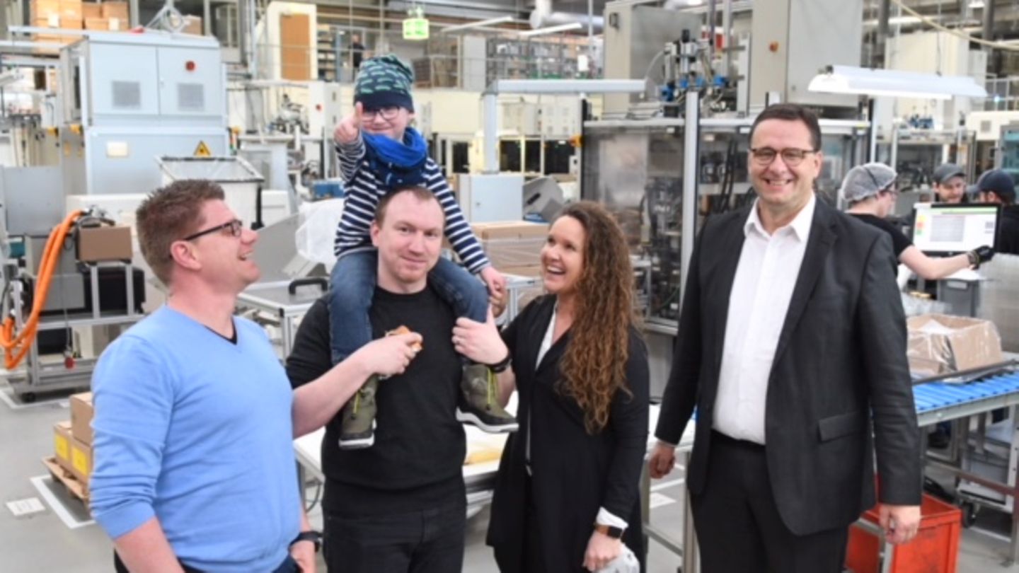Drei Männer und eine Frau stehen lachend in einer Fabrik. Ein Mann trägt ein Kind auf der Schulter, das den Daumen hoch zeigt.
