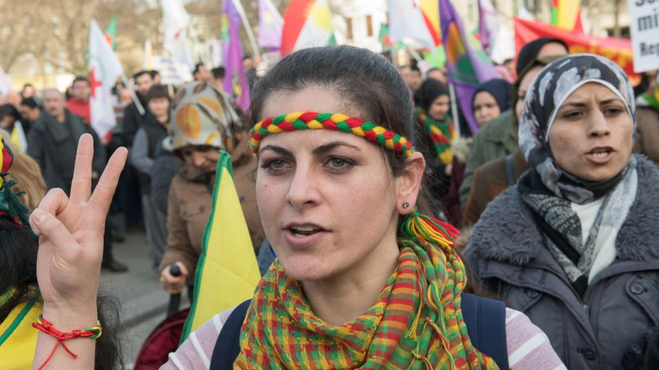 Eine Demonstrantin mit gelb-rot-grünem Stirnband und Halstuch zeigt mit ihrer rechten Hand ein Peace-Zeichen
