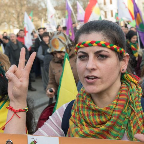Eine Demonstrantin mit gelb-rot-grünem Stirnband und Halstuch zeigt mit ihrer rechten Hand ein Peace-Zeichen