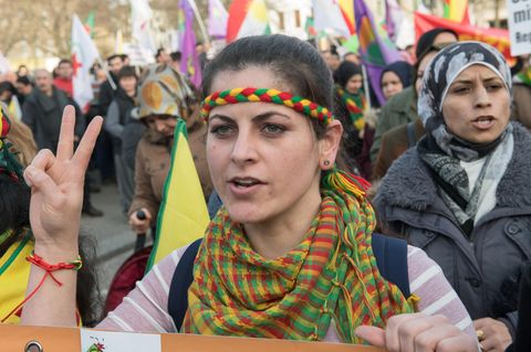 Eine Demonstrantin mit gelb-rot-grünem Stirnband und Halstuch zeigt mit ihrer rechten Hand ein Peace-Zeichen