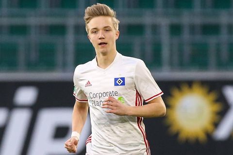 Ein junger Mann mit dunkelblonden, hochgeföhnten Haaren läuft in weißem HSV-Trikot und roter Hose über einen Fußballplatz