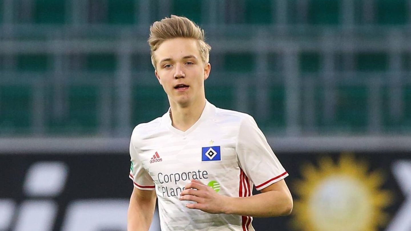 Ein junger Mann mit dunkelblonden, hochgeföhnten Haaren läuft in weißem HSV-Trikot und roter Hose über einen Fußballplatz