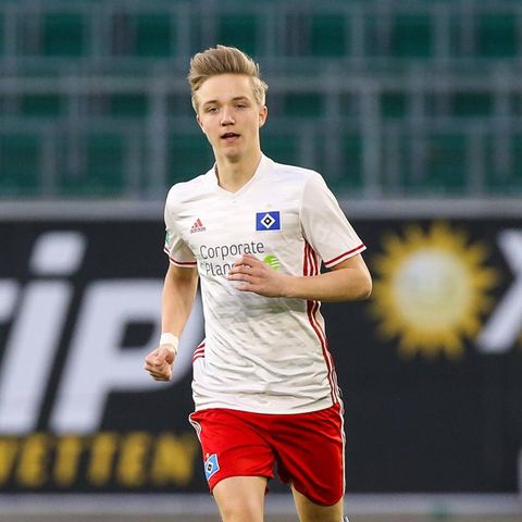 Ein junger Mann mit dunkelblonden, hochgeföhnten Haaren läuft in weißem HSV-Trikot und roter Hose über einen Fußballplatz